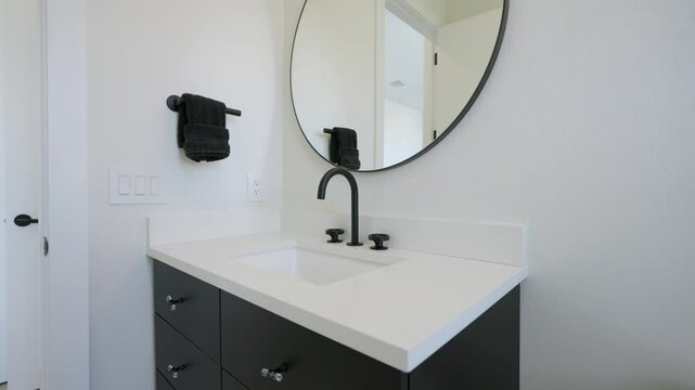 Swellish bathroom sink area with countertop, black faucet and handles. Round mirror with black frame. Black towel hanging on wall-mounted holder. Vanity with silver knobs