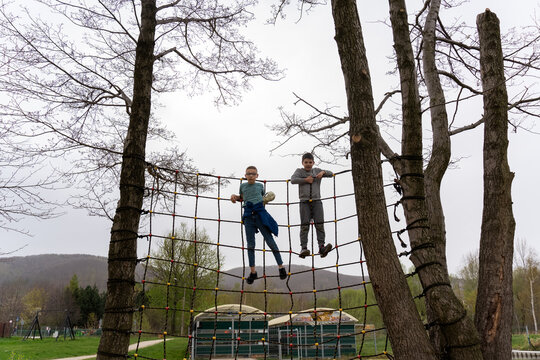 Two boys climbing on rope net obstacle course between trees on cloudy day in the park. Concept of outdoor adventure, teamwork, and active childhood fun in nature. High quality photo