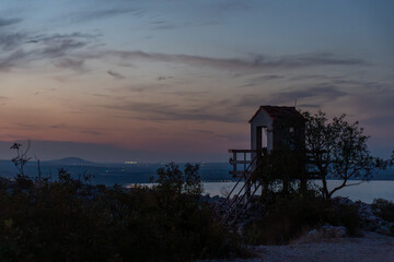 Scenic sunset view with small rustic observation tower surrounded by trees overlooking calm landscape with distant lights and hills. Concept of tranquility, countryside beauty, and peaceful nature