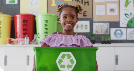 Happy african american schoolgirl standing in classroom, smiling, holding recycling bin