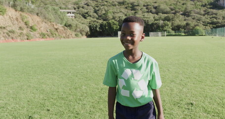 An African American boy wearing a green recycling t-shirt stands on a sunny soccer field, with copy