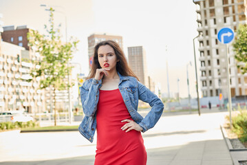 Fototapeta premium A woman in a red dress is walking in the city. She is wearing a denim jacket.