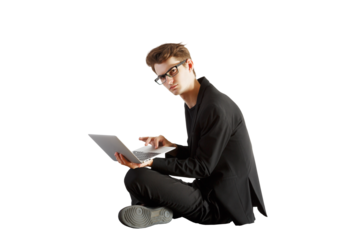 Young man in glasses wearing a formal outfit seated on the floor while using a laptop, isolated on a white background, concept of work