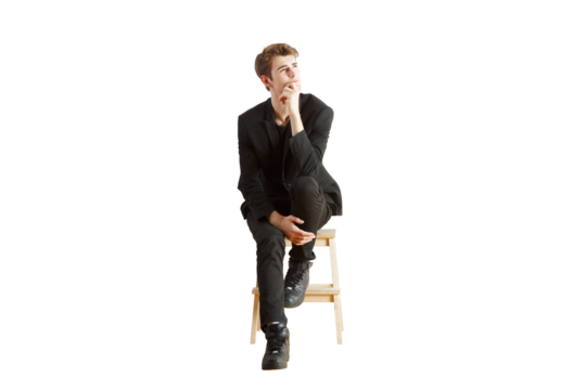 Young man in asuit sitting on a wooden stool, posing thoughtfully, isolated on a white background. Concept of contemplation and fashion