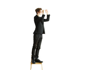 Obraz premium Full-length image of a man in asuit standing on a wooden chair, reaching upward, against a white background. Concept of effort or problem-solving