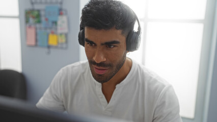 Hispanic man working indoors wearing headphones in an office setting, focused on a task with a...