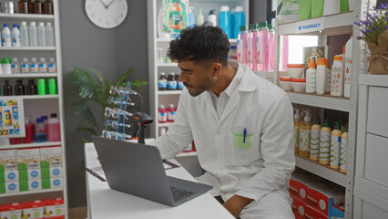 Young hispanic man working on a laptop in a well-organized pharmacy filled with various medical and beauty products