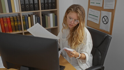 Woman working in an office, examining documents at her desk, with blonde hair and glasses, in a...