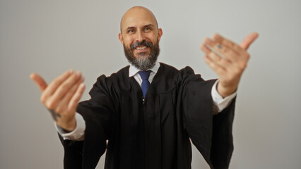Handsome hispanic man with a beard and bald head, wearing a gown, making a come-hither gesture with his hands, isolated against a white background wall.