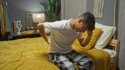 A young, handsome hispanic man sitting on a bed in a bedroom, showing signs of discomfort, wearing glasses and pajamas, captured in an indoor home setting with decor and furniture.