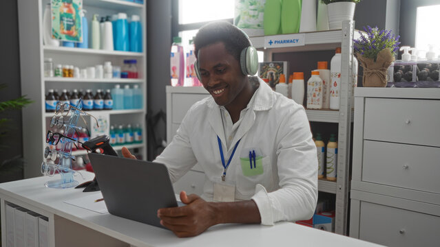 Handsome young african american man working in a pharmacy store wearing a lab coat, smiling while using a tablet and wearing headphones with shelves of medications and products behind him.
