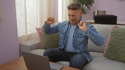 Middle-age man celebrating success at home, sitting on a cozy couch in a brightly lit living room,...