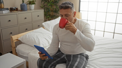 Middle-aged man drinking coffee and using tablet in cozy home bedroom, creating a relaxed indoor setting