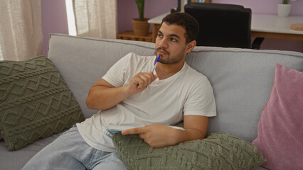 Fototapeta premium Young man thinking while sitting on a couch in a cozy living room, holding a pen against his lips in a pensive mood.