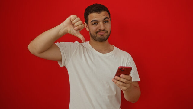 Young man holding smartphone with thumbs down gesture over isolated red background