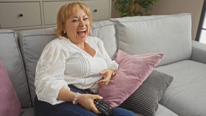Obraz premium A beautiful elderly caucasian woman laughs joyfully while sitting on a sofa in a cozy living room, holding a remote control in her hand.