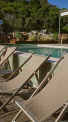 Empty deck chairs by an outdoor swimming pool set against lush greenery and sunny skies, perfect for relaxation and leisure in a serene natural setting.