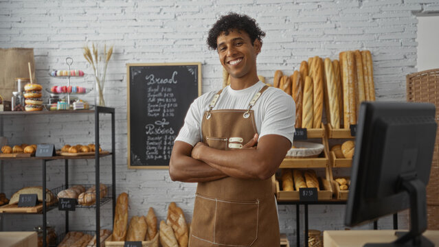 Young man with arms crossed smiling in a bakery displaying various bread and pastries with a chalkboard menu in the background