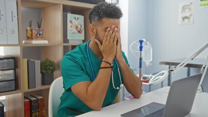 Portrait of a young hispanic male vet in a veterinary clinic, appearing stressed while sitting at a desk with his hands covering his face.