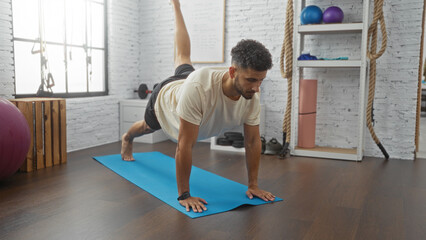 Obraz premium Handsome young hispanic man performing yoga on a mat in a gym, showcasing athleticism and concentration in a modern indoor sport center setting.