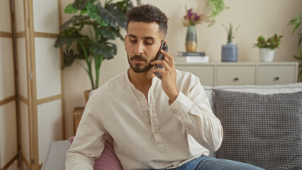 Young hispanic man having a phone call while sitting in a stylish living room at home.