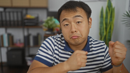 Fototapeta premium A young chinese man in a striped polo shirt poses with a playful expression in a modern office, with shelves and plants in the background, showcasing an attractive indoor professional environment.