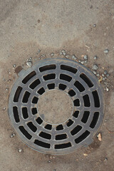 Detailed Image Of A Circular Metal Drain Cover Situated On A Gritty Urban Pavement, Capturing The Texture Of The Ground And The Unique Design Of The Cover.