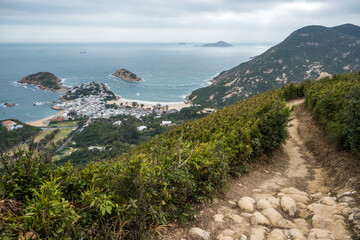 Dragons Back Hike, Shek O Peak, Hong Kong: A winding dirt path on a green mountainside overlooks a coastal village nestled between rocky headlands.