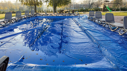 High-angle view of a swimming pool covered with a blue tarp, surrounded by empty lounge chairs,...