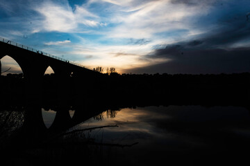 Silhouette of an Arched Bridge at Sunset Reflecting on Calm Waters in Fuentes Claras, Avila, CyL