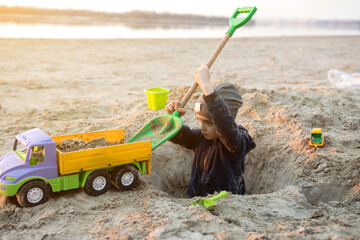A child playing on the beach with a toy truck and shovel, digging a hole in the sand. The scene captures the joy and creativity of outdoor playtime by the water.