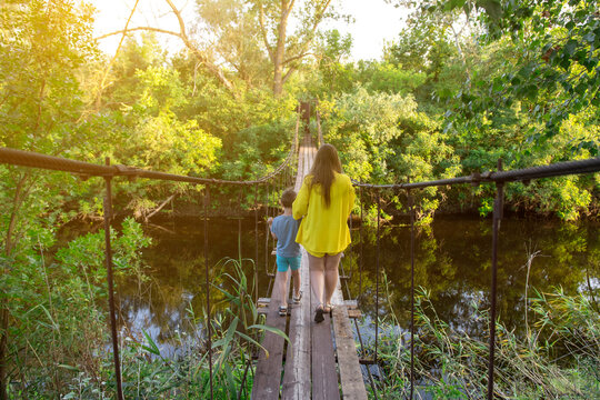 Mother and son crossing a rustic wooden bridge in a dense forest, surrounded by vibrant greenery and a serene river. Ideal for concepts of bonding, exploration, and outdoor activities