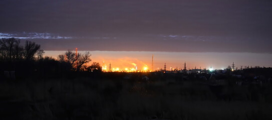 An evening landscape with city lights and the lights of a working factory illuminating the horizon under a dark twilight sky, creating a serene and atmospheric scene.