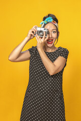 Emotional Asian girl dressed in pin-up style with vintage camera in her hands doing photo shoot on bright yellow background. Isolated.