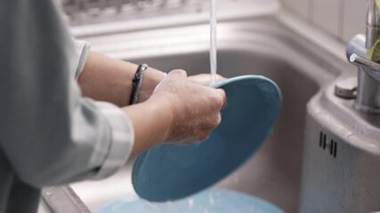 Close-up of hands washing a blue plate under running water in the kitchen sink. The moment captures everyday household chores with attention to cleanliness and care.