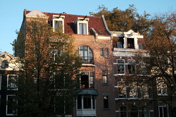 Fototapeta premium Fassaden schöner alter Wohnhäuser mit Mansarde, Dachgaube und alten Bäumen im Herbst im Licht der Abendsonne an der Keizersgracht in der Altstadt von Amsterdam, der Hauptstadt der Niederlande