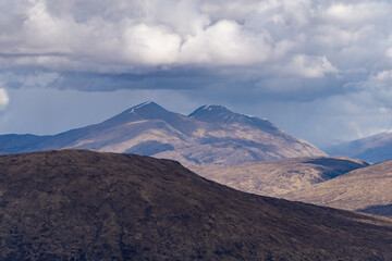 This image features rugged, sunlit mountain peaks with patches of snow, surrounded by shadowed hills under a dramatic sky of thick, brooding clouds, creating a striking and moody scene.