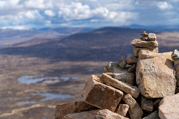This image shows a stone cairn in the foreground with a vast, rugged landscape of peat bogs, hills,...