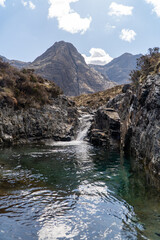 This image captures a serene mountain pool with crystal-clear water and a small waterfall framed by rocky terrain. In the background, towering, rugged mountains rise against a partly cloudy sky.