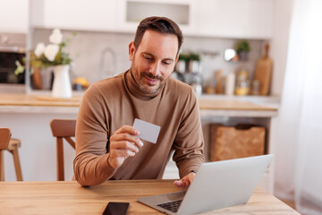 Mid adult man doing online shopping over laptop and paying with credit card on dining table at home