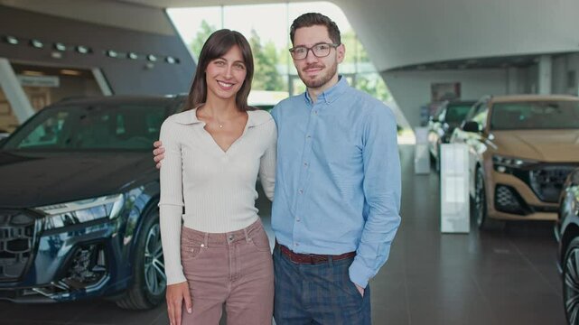 Caucasian couple spending time in car salon. Handsome man and woman standing next to several cars in background. Pretty woman with natural makeup hugging her husband. People smiling to camera.