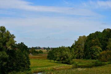 A view of a shallow yet vast pond or lake flowing next to vast fields covered with herbs, reeds, and various crops located next to some tiny forests and moors spotted on a cloudy summer day in Poland