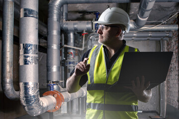 Man engineer in basement. Worker stands with laptop near heating pipes. Dark basement with boiler...