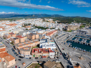 Aerial perspective of Tarifa, Spain, highlighting Guzman Castle, a marina with boats, whitewashed buildings, rolling hills, and wind turbines.