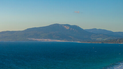 Fototapeta premium The image captures an aerial view of Tarifa, Spain, featuring the blue sea, distant mountain range, and kite surfers with colorful kites along the shoreline.
