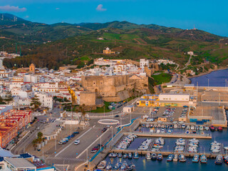 The image captures an aerial view of Tarifa, Spain, highlighting Guzman Castle, whitewashed buildings, lush green hills, and a bustling harbor with boats.