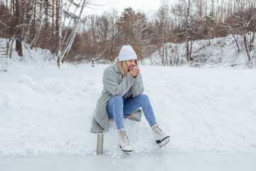 Fototapeta premium Lovely young woman relaxing after riding ice skates and drinking hot drink from thermo pot on the ice rink. Girl sitting on snow and smiling.