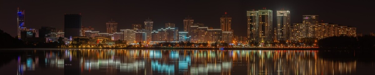 night city. widescreen panoramic view of a modern quarter with tall buildings on the coast of a large lake with electric lighting and reflection on the water surface. Minsk, Belarus. side view 25x5