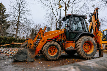 Yellow backhoe loader parked at a muddy construction site surrounded by autumn trees. High quality...
