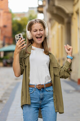 Smiling happy young woman in wireless headphones dancing, listening favorite energetic disco music singing holds smartphone celebrate win outdoors. Girl standing on urban city street. Town lifestyles.